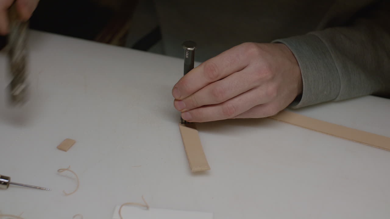 An artisan carefully punches holes in a leather strip using a hammer and chisel on a white work surface.