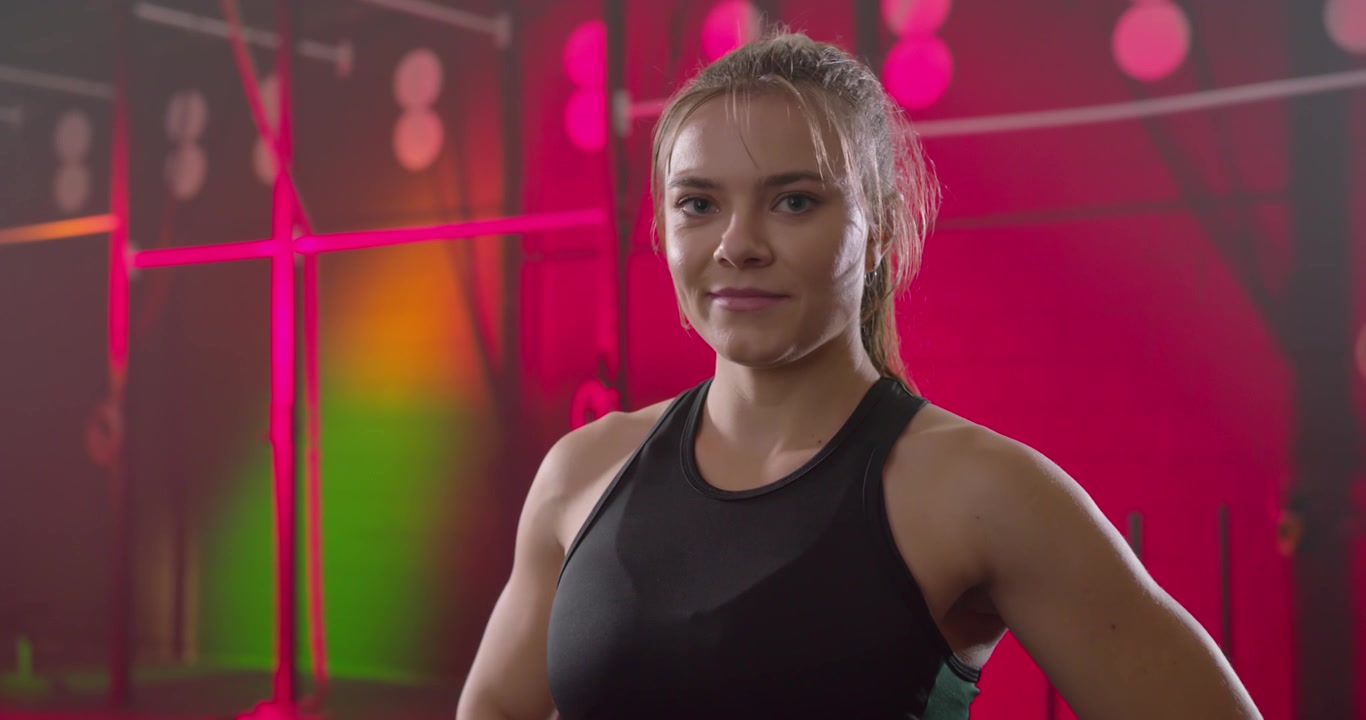 Atleta feminina usando top esportivo posando enquanto a câmera se aproxima do equipamento, suavizando o sorriso para o anúncio