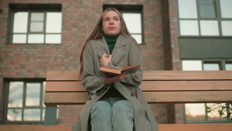 lady seated on wooden bench holding book on lap, adjusting coat while feeling cold, wearing grey jacket and jeans against modern brick building background
