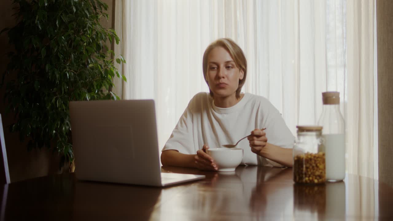 Woman eating cereal while working on laptop at home
