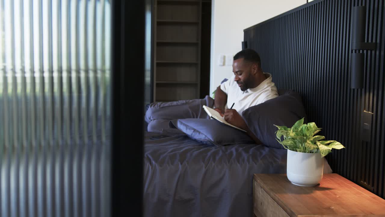 African American man sitting on bed writing in notebook with pen while frosted panel shifting