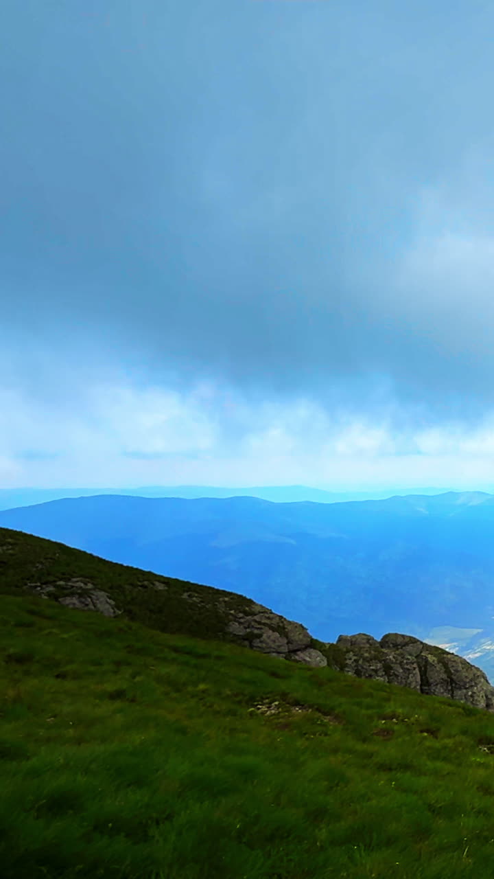 Il primo piano presenta un crinale roccioso scuro e erba verde che si affacciano su vasti strati di sagome blu di montagne sotto un cielo nuvoloso. Cima di montagna rumena. Video verticale.