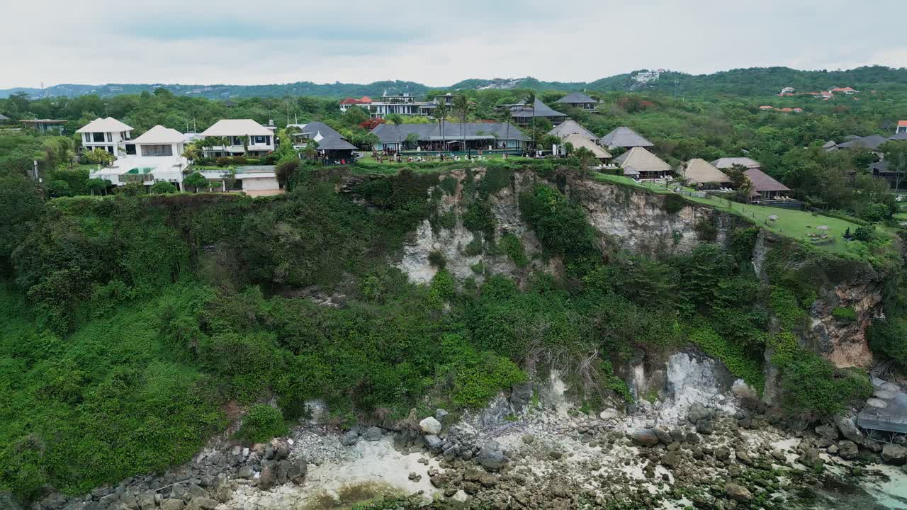Uluwatu features a breathtaking coastal landscape where towering cliffs meet deep blue water and luxury villas sit along the edge offering stunning aerial drone views of Bali's wild southern shore.