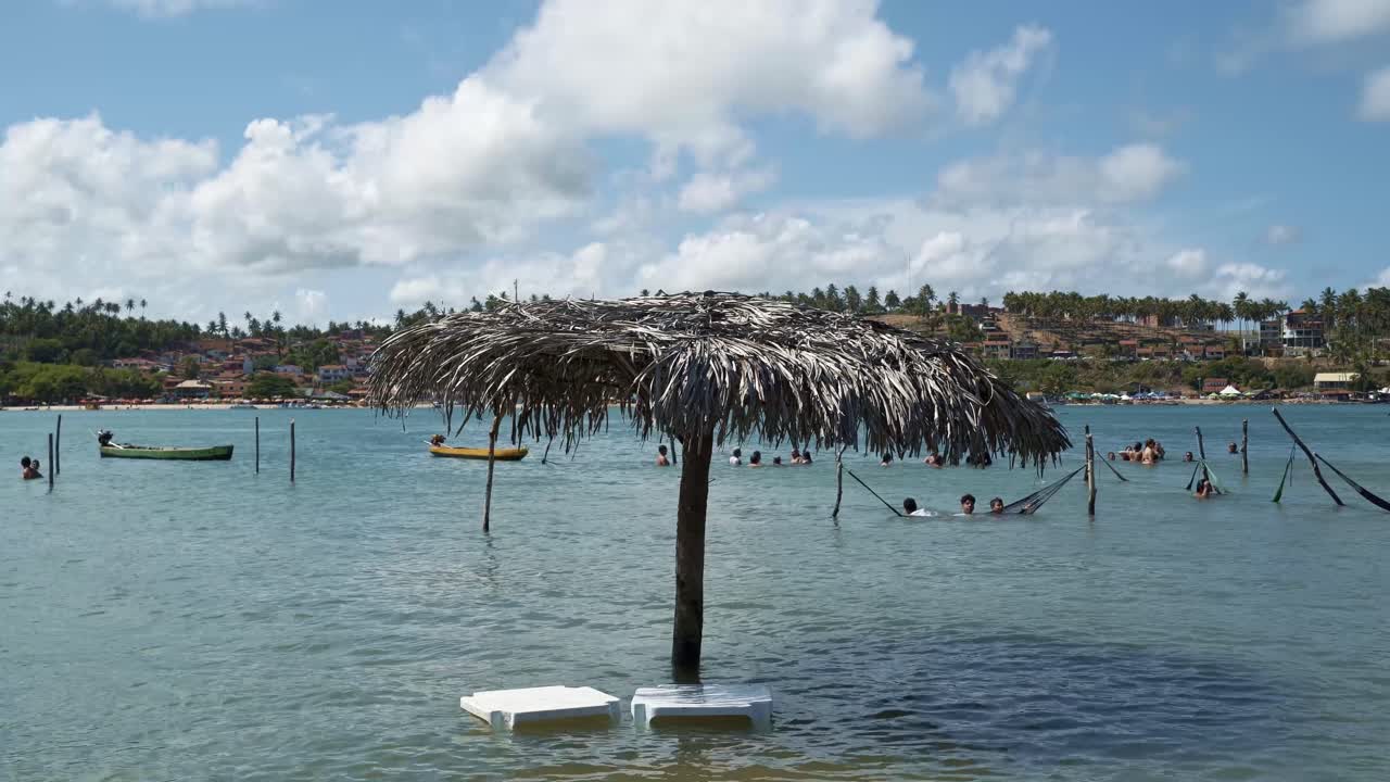 una sombrilla de paja de palma hecha a mano y mesas de plástico sumergidas por un río turquesa tropical durante la marea alta en la playa de restinga frente a la barra de cunhaú, rio grande do norte, brasil