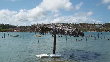 A-hand-made-palm-thatch-umbrella-and-plastic-tables-being-submerged-by-a-tropical-turquoise-river-during-high-tide-on-Restinga-beach-across-from-the-Barra-de-Cunhaú,-Rio-Grande-do-Norte,-Brazil