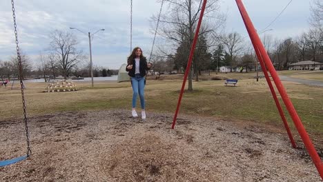 slow motion footage of a beautiful brunette college teenager swinging in a city park wearing jeans, a white shirt and a black jacket