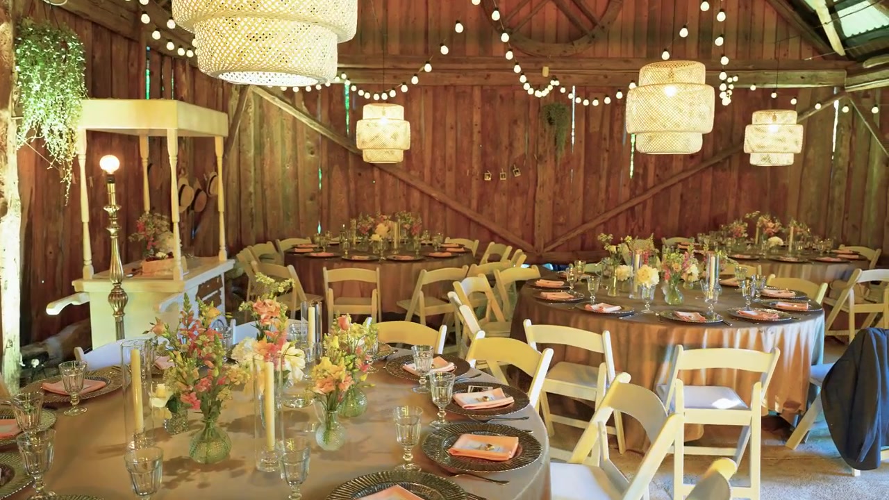 Charming rustic barn wedding reception features round tables with floral centerpieces, elegant place settings, string lights, and lanterns. Warm ambient lighting creates a cozy, festive atmosphere.