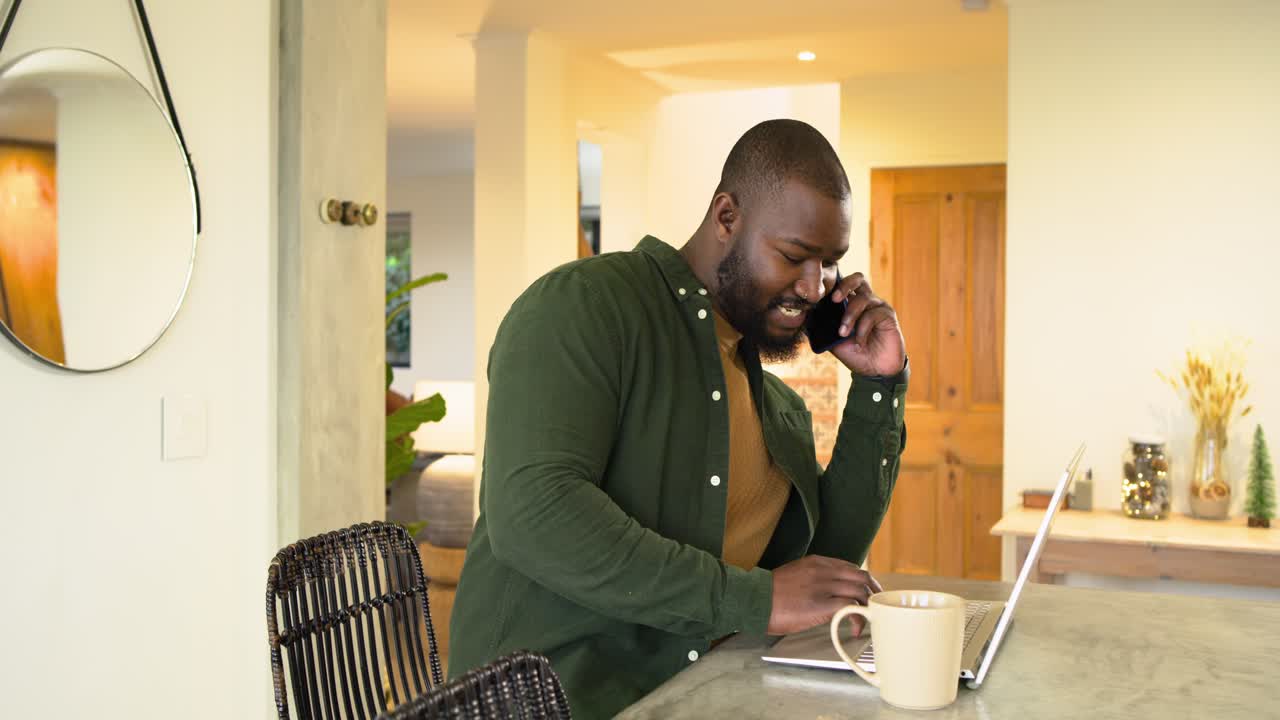 In kitchen African American man receiving call greeting smartphone caller and typing on laptop