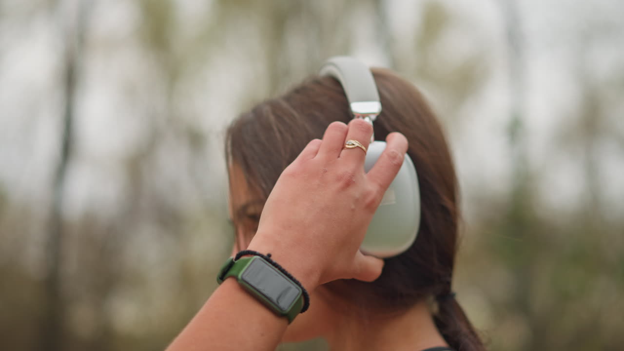 Side view of a young lady wearing headphones, focused and adjusting the volume on her headphones, with blurred natural background, perfect for videos about music, tech, or concentration