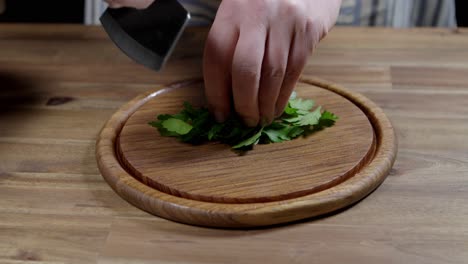 chef rolling fresh parsley to finely chop with kitchen ax knife on round wooden board