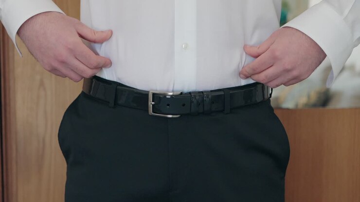 Primo piano dello sposo che stringe una cintura di pelle nera sopra una camicia bianca e pantaloni scuri dell'abito.