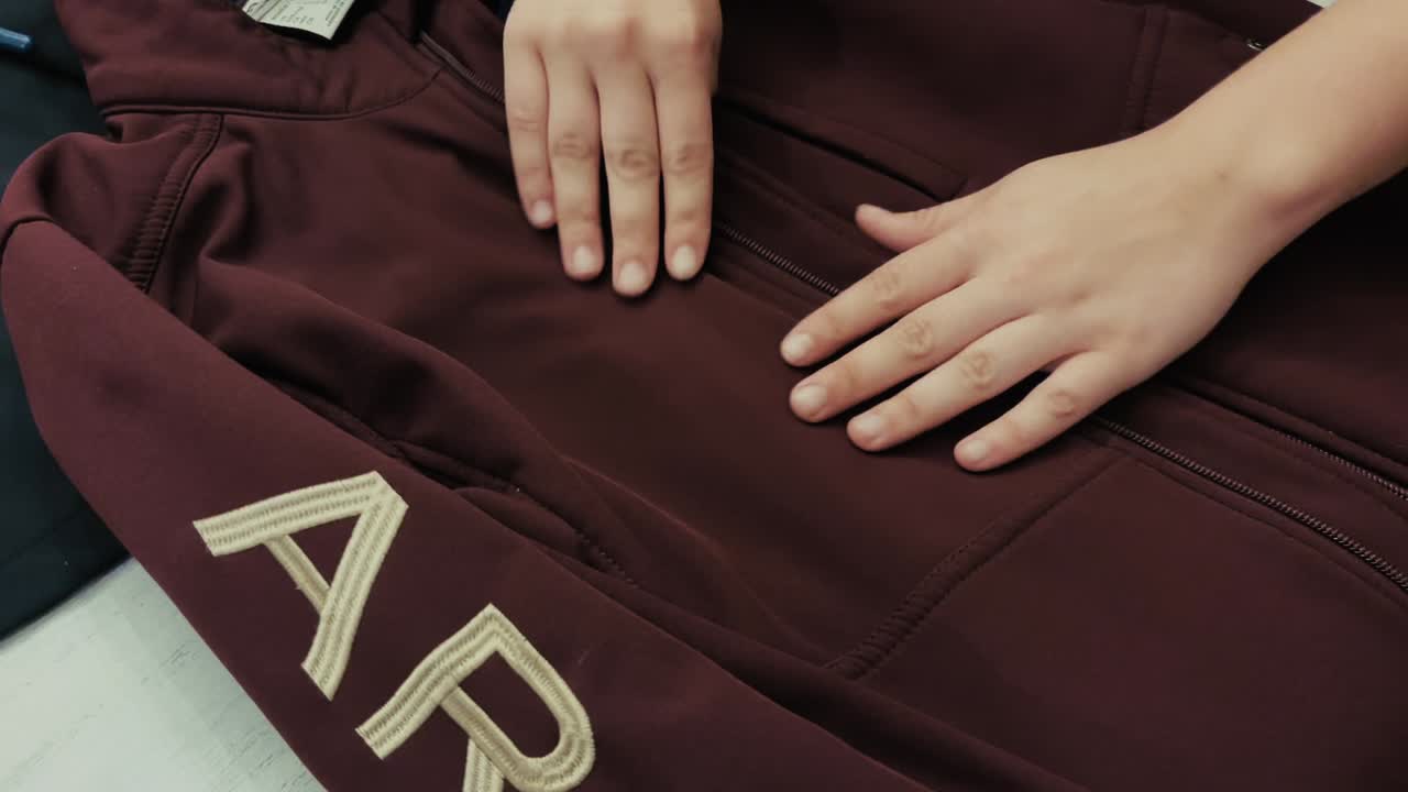 Close-up of two hands smoothing the surface of a maroon jacket laid flat, showing fabric texture and stitching details