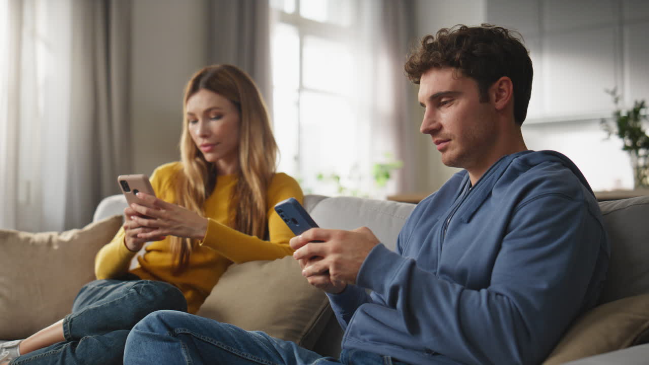 Two lovers messaging smartphones at home weekend. Serious duet sitting sofa