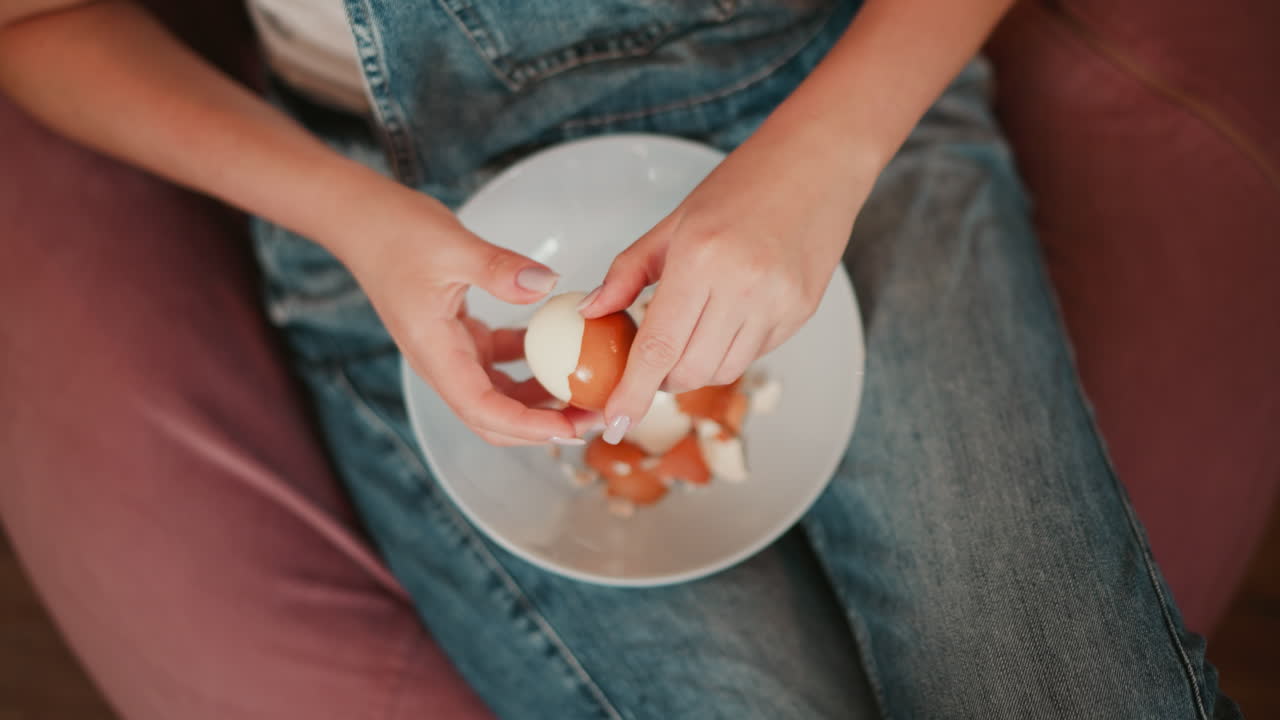 Close up aerial view of person seated on pink bean bag gently peeling shell off brown boiled egg over white bowl, broken shells scattered, casual setting in denim overalls