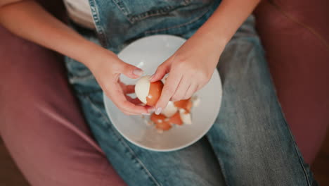 close up aerial view of person seated on pink bean bag gently peeling shell off brown boiled egg over white bowl, broken shells scattered, casual setting in denim overalls