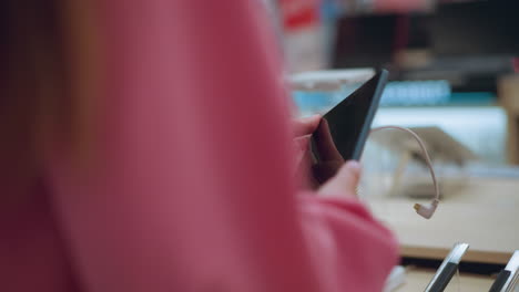 lady seen from behind picking up a tablet from its stand in a store, tablet is connected with a security cord, while other tablets are arranged neatly on the display, she closely inspects it
