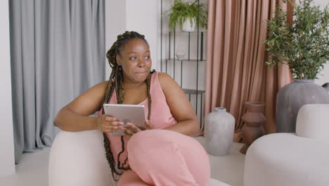 woman with braids sitting on a big sofa while using a tablet, then looks at a side