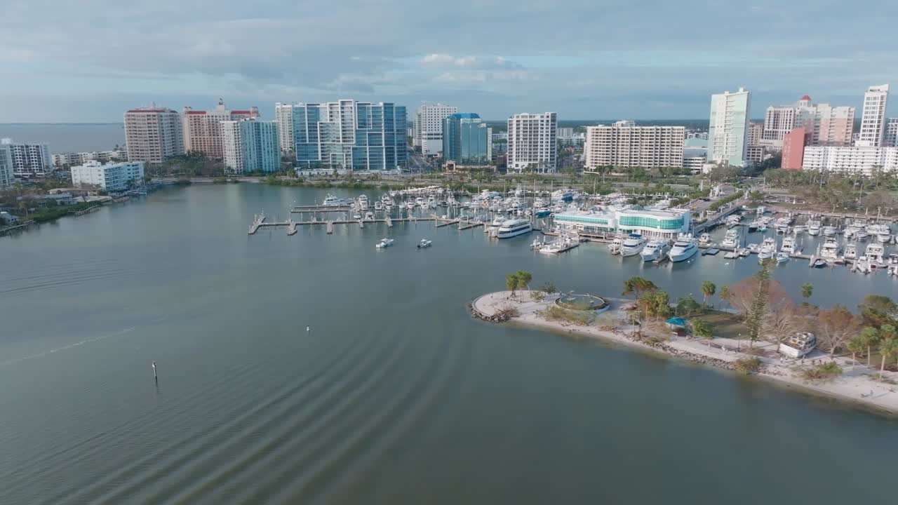 Aerial flight over Sarasota Bay, Marina Jack, and downtown Sarasota, showing debris, grounded boats, and downed trees post-Hurricane Milton. Small boats approach marina, with waves and bystanders.