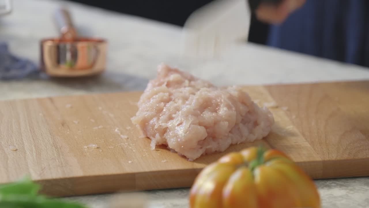 Minced raw chicken is being finely chopped with a ceramic knife on a wooden cutting board, with blurred vegetables and kitchen tools in the soft-lit background.
