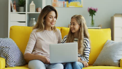 Fröhliche,-Gut-Aussehende-Mutter-Und-Tochter-Sitzen-Zusammen-Auf-Der-Gelben-Couch-Und-Unterhalten-Sich-Auf-Dem-Laptop-Im-Gemütlichen-Wohnzimmer.-Drinnen.