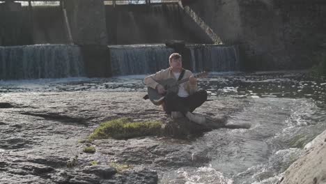 young man playing guitar sitting on the bank of a mountain river on a background of rocks. concept of freedom relaxation. place