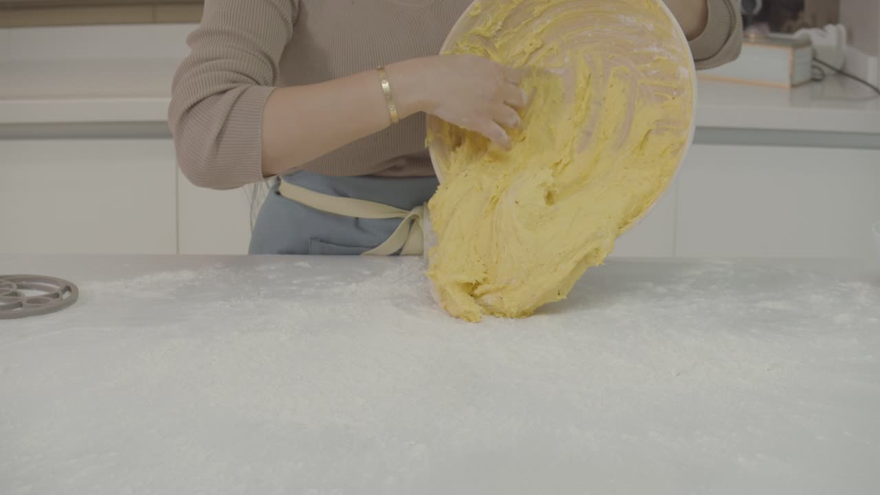 Close up view of a lady pouring a sponge dough from a bowl onto a white table before baking in the kitchen.