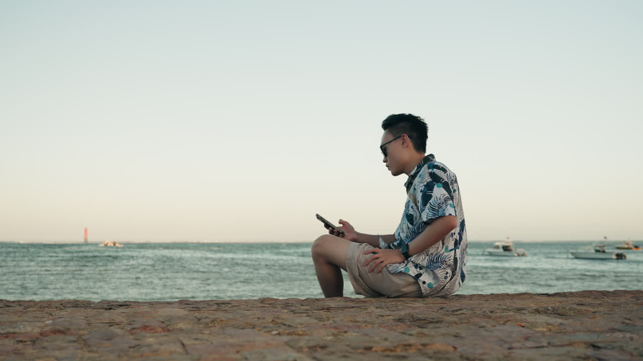 아시아 남자 휴가 휴가 해변에서 휴대폰을 잡고 선글라스를 입고 앉습니다.