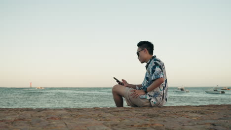 hombre asiático sostener el teléfono en la playa en vacaciones vacaciones sentarse con gafas de sol
