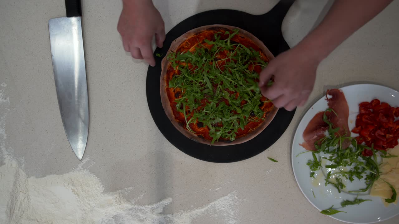 Top down view of a chef making pizza with various condiments. Overhead view of cook adding arugula on a pizza dough with ketchup