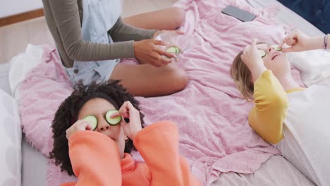 felices y diversas adolescentes amigas acostadas en la cama con rodajas de pepino en la cara, cámara lenta