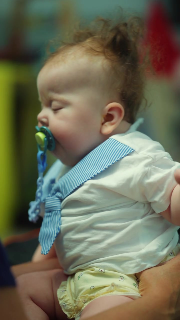 Baby boy sleeping peacefully in parents arms, wearing a white shirt with bow tie and pacifier during a nap