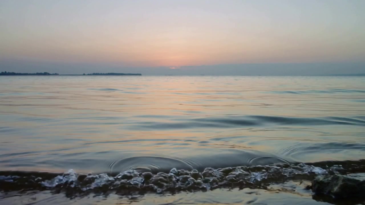 Il sole che tramonta proietta fasce pallide sul mare calmo al tramonto, con il promontorio sinistro e le rocce basse che schiumano.