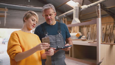 Female-Apprentice-Learning-From-Mature-Male-Carpenter-With-Digital-Tablet-In-Furniture-Workshop