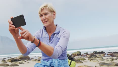 mujer excursionista senior con mochila tomando fotos usando un teléfono inteligente mientras está sentada en la roca