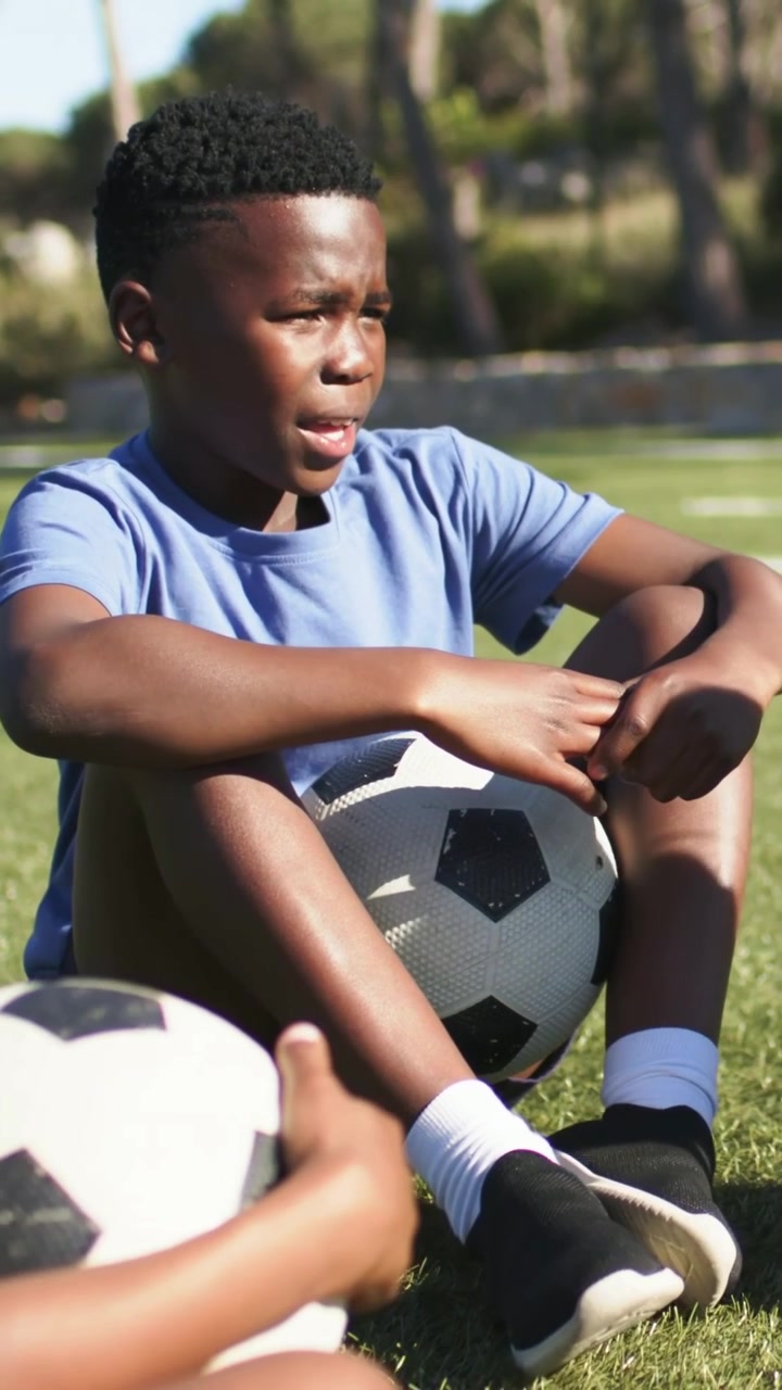 Vídeo vertical: Jovem de azul sentado no campo, observando amigo trazer a bola, com bola preta ou branca, ouvindo o treinador.
