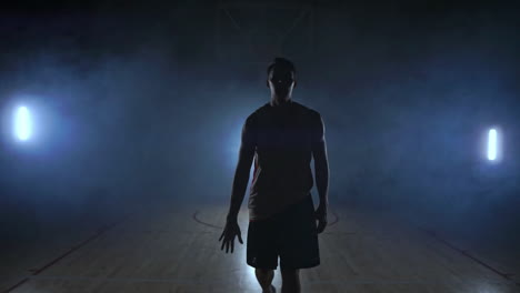 basketball player goes straight to the camera in a dark room with a backlit back in the smoke looking at the camera in slow motion. steadicam