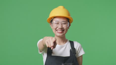 close up of asian woman worker wearing goggles and safety helmet smiling and touching her chest then pointing at you while standing in the green screen background studio