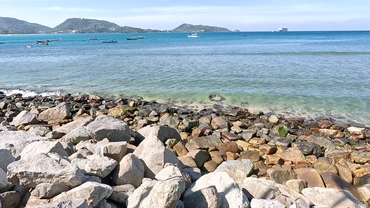 Calm ocean waves gently lap against rocky shores under bright sunlight at Kalim Beach, Phuket, with distant hills in view
