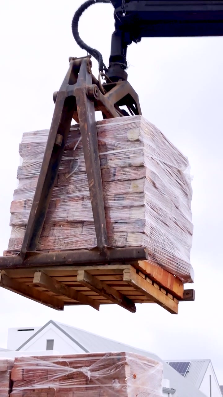 Gru caricatrice che utilizza una pinza per mattoni per sollevare un pallet di mattoni da un camion da trasporto. Verticale.