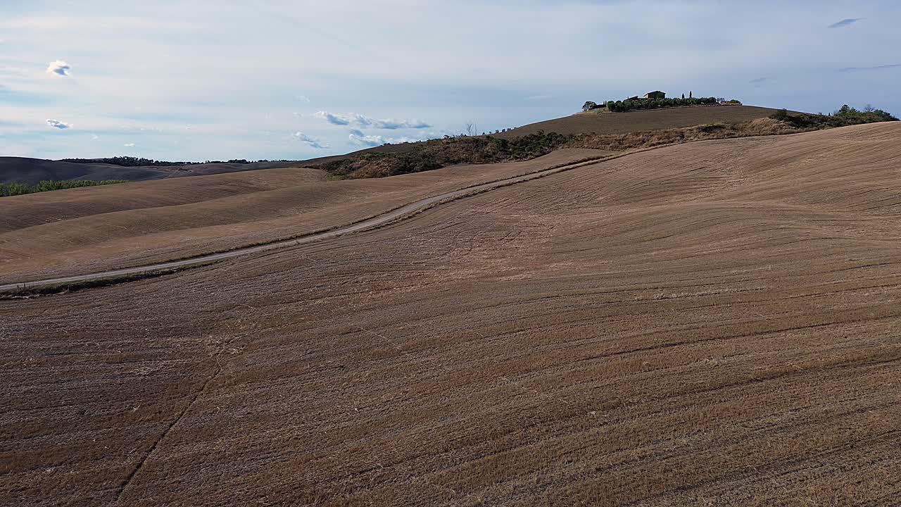 Winding dirt road cutting through Tuscany's golden rolling hills under a vast blue sky with clouds.
