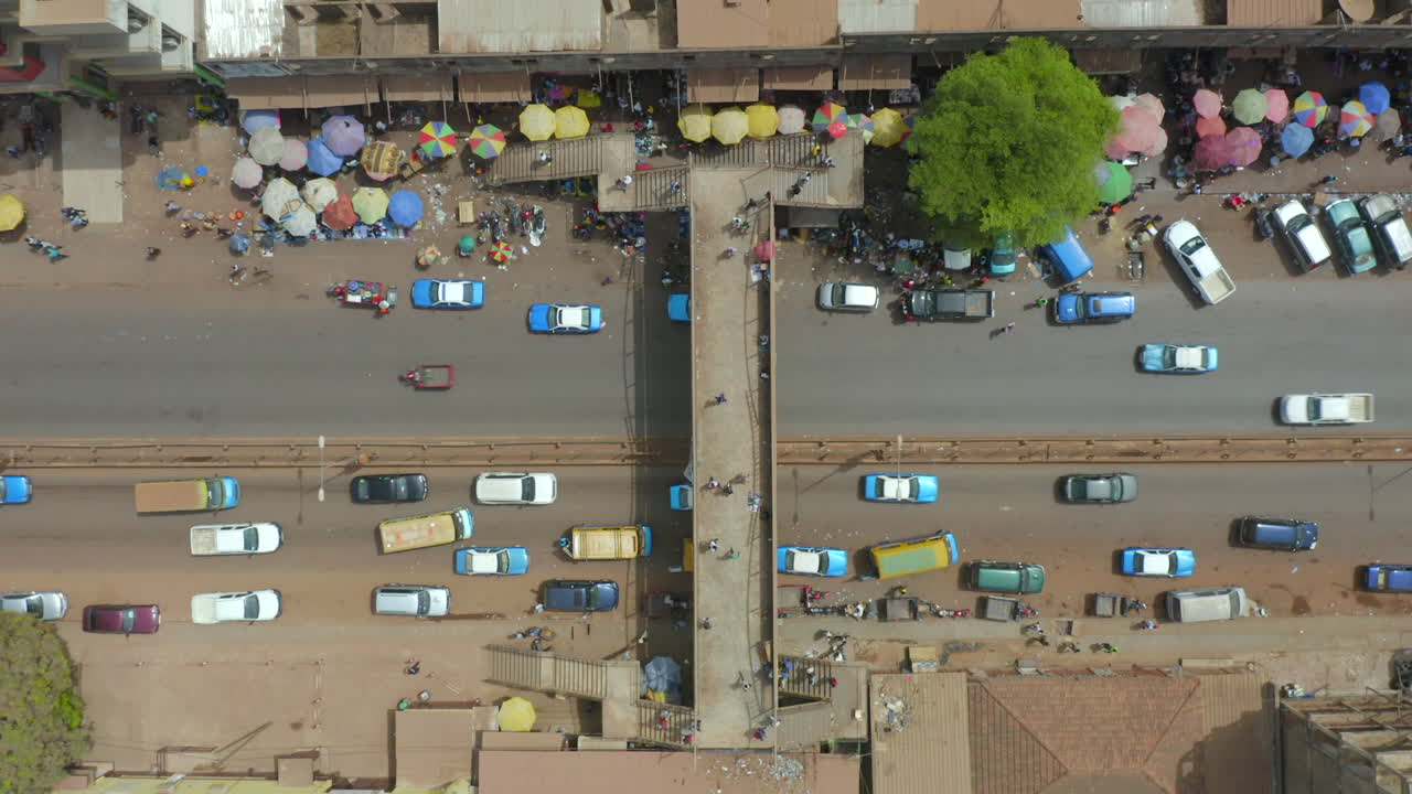 A high-angle drone shot directly over a busy street in Guinea-Bissau, West Africa. Features an overpass, traffic congestion, and colorful market stalls, emphasizing movement and dense urban life