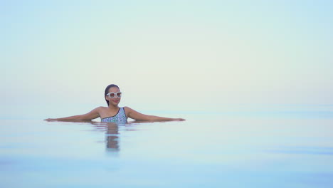 A-woman-with-her-arms-spread-out-along-the-edge-of-an-infinity-pool-appears-to-be-floating-in-space-against-an-ocean-horizon