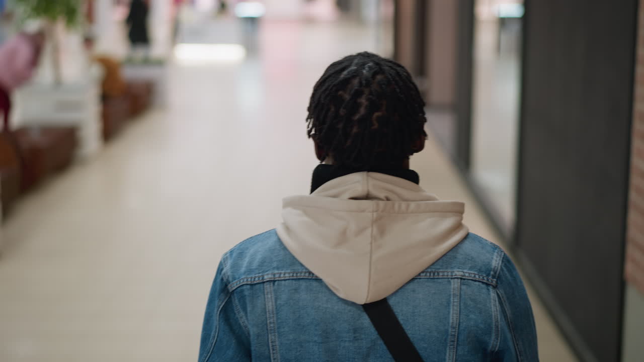 back view of young man wearing beige hoodie under denim jacket walking through bright modern mall corridor with dreads hairstyle blurred storefronts brick wall ceiling lights overhead