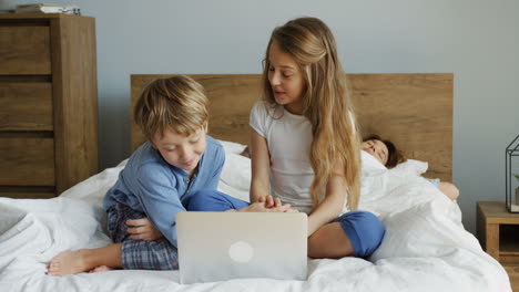 lindos niños pequeños acostados frente a la cámara en la cama y usando una computadora portátil mientras sus padres yacen detrás de ellos por la mañana