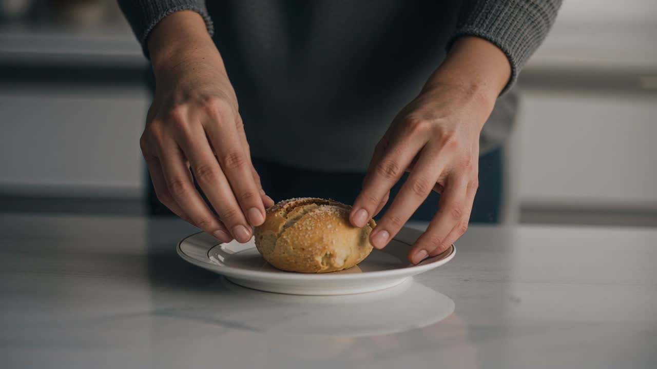 Hands reaching nudging, rotating bread roll onto white plate on counter, centering in grey sweater