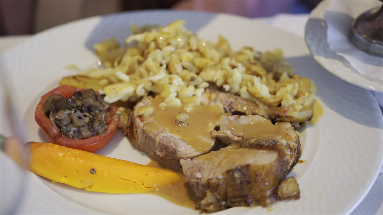Close up of a server arranging a portion of egg noodle near a few pieces of pork, cooked carrot and stuffed tomato on a white plate
