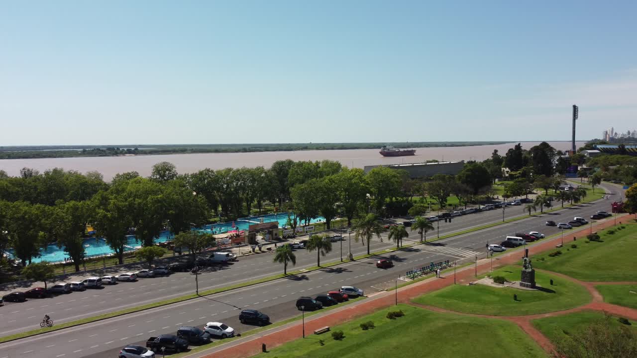 A drone captures an orbital view of the Paraná River in Rosario, showing a pool complex and a busy avenue with numerous cars, creating a dynamic urban scene.