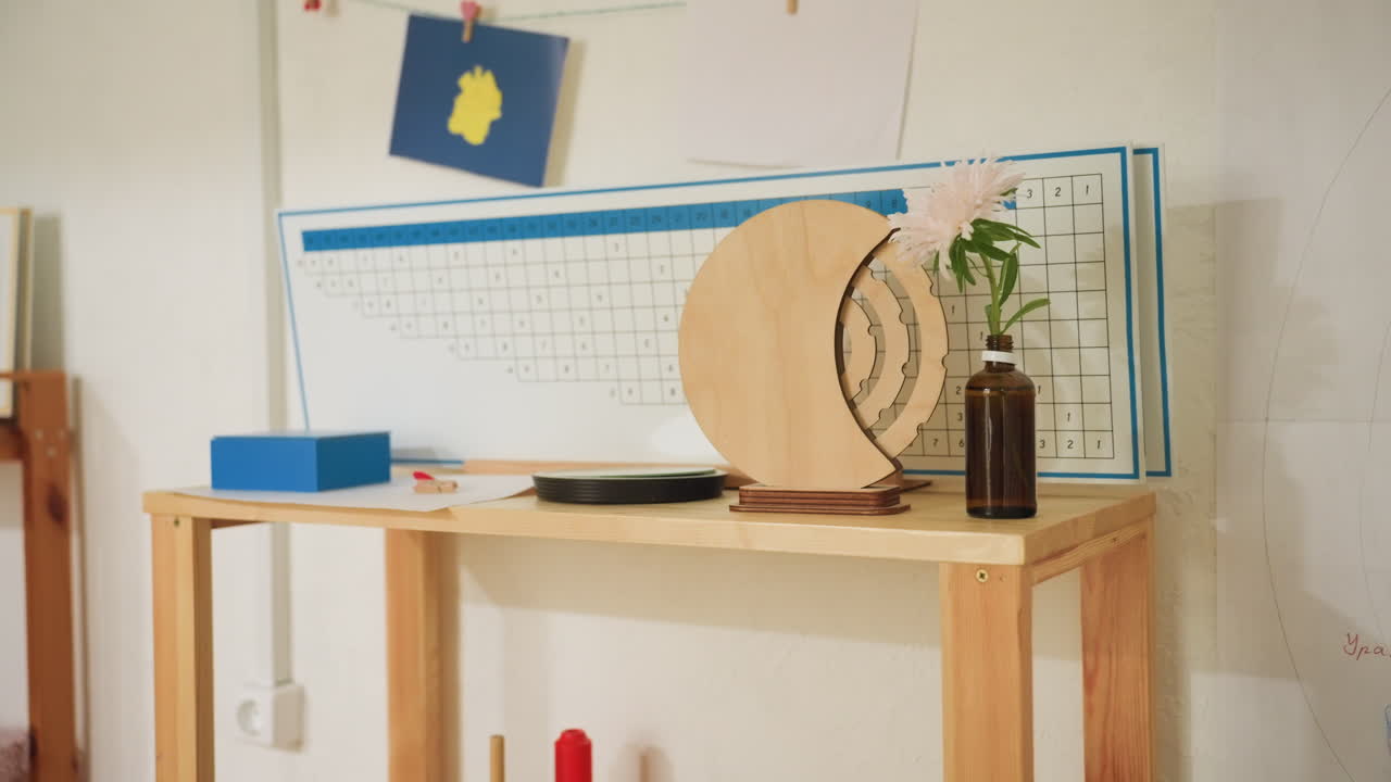 Wooden shelf filled with Montessori learning tools including geometric blocks, tactile frames, and sorting boxes. Decorative flower in glass bottle and educational chart