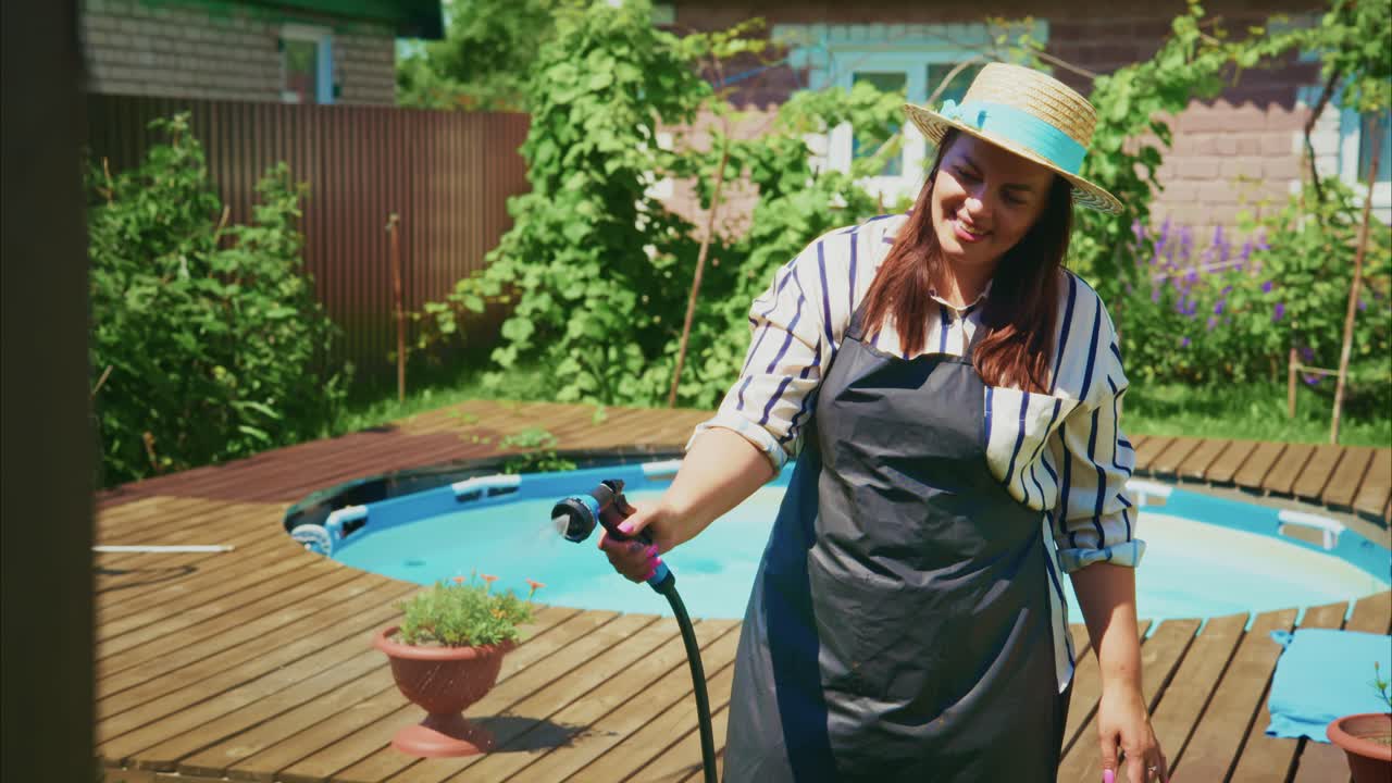 Woman in apron watering plants near swimming pool in sunny backyard garden setting