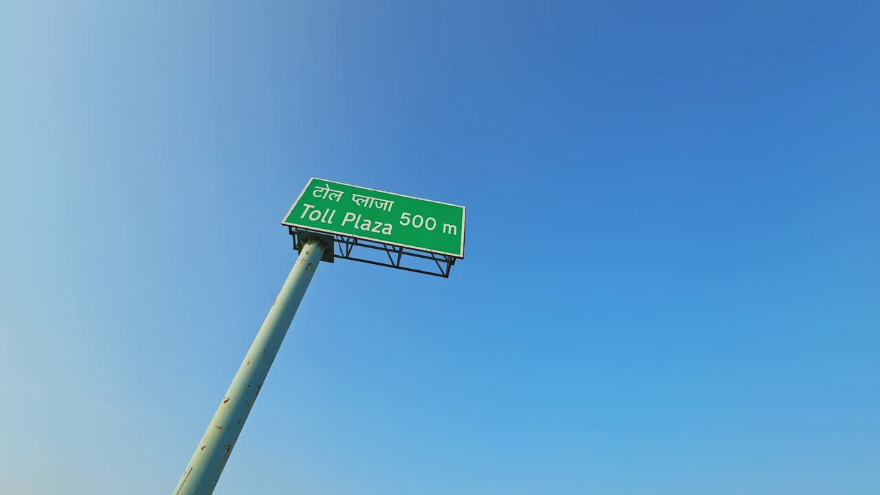 Dramatic low-angle view from directly below a green “Toll Plaza 500 m” highway sign in Hindi and English, mounted on a tall pole against a clear blue sky, captured with a subtle orbiting motion